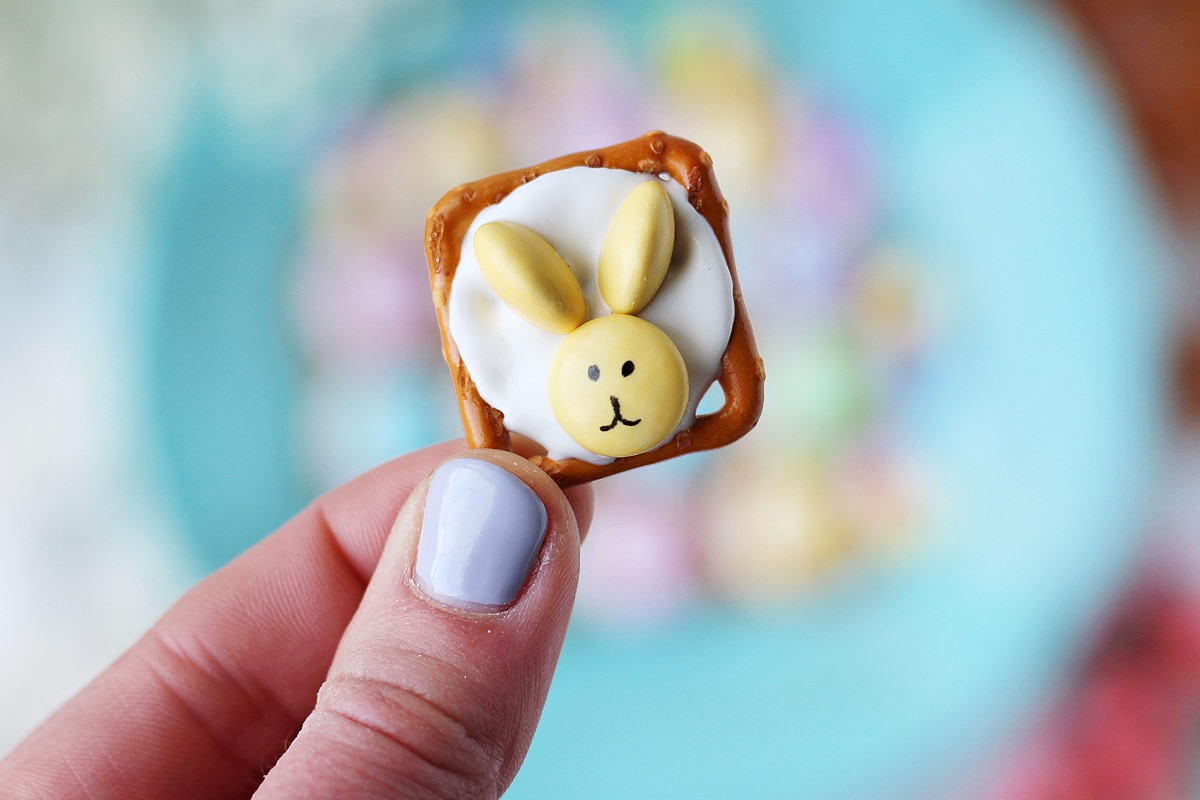 A woman's fingers holding up a Easter bunny pretzel with a pastel yellow M&M.
