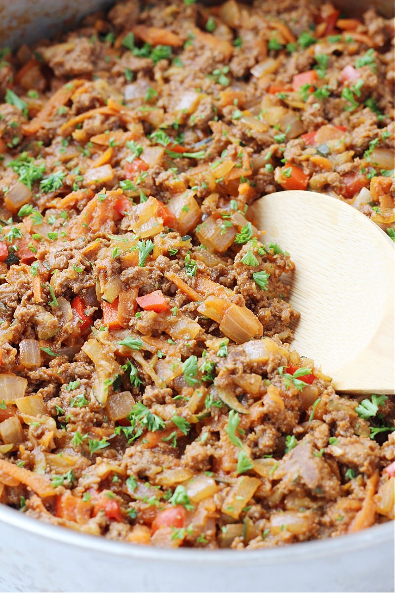 A wooden spoon digging into a pan of sloppy joes with meat and fresh veggies.