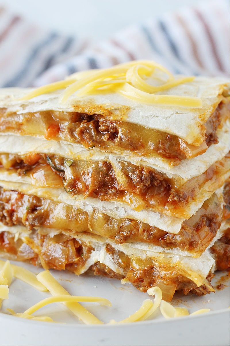 A stack of sloppy joe quesadilla wedges on a white plate topped with shredded cheese.