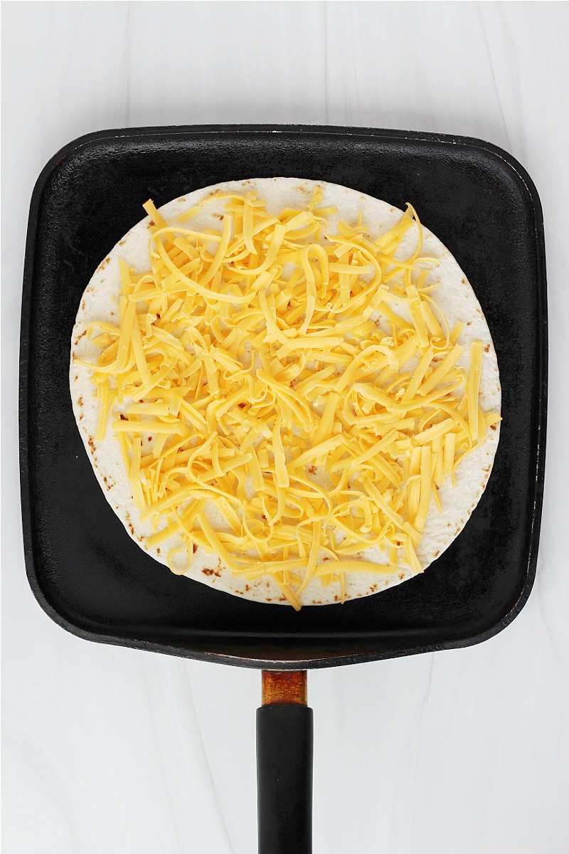 Overhead photo of a tortilla with shredded cheese on top on a black pan.