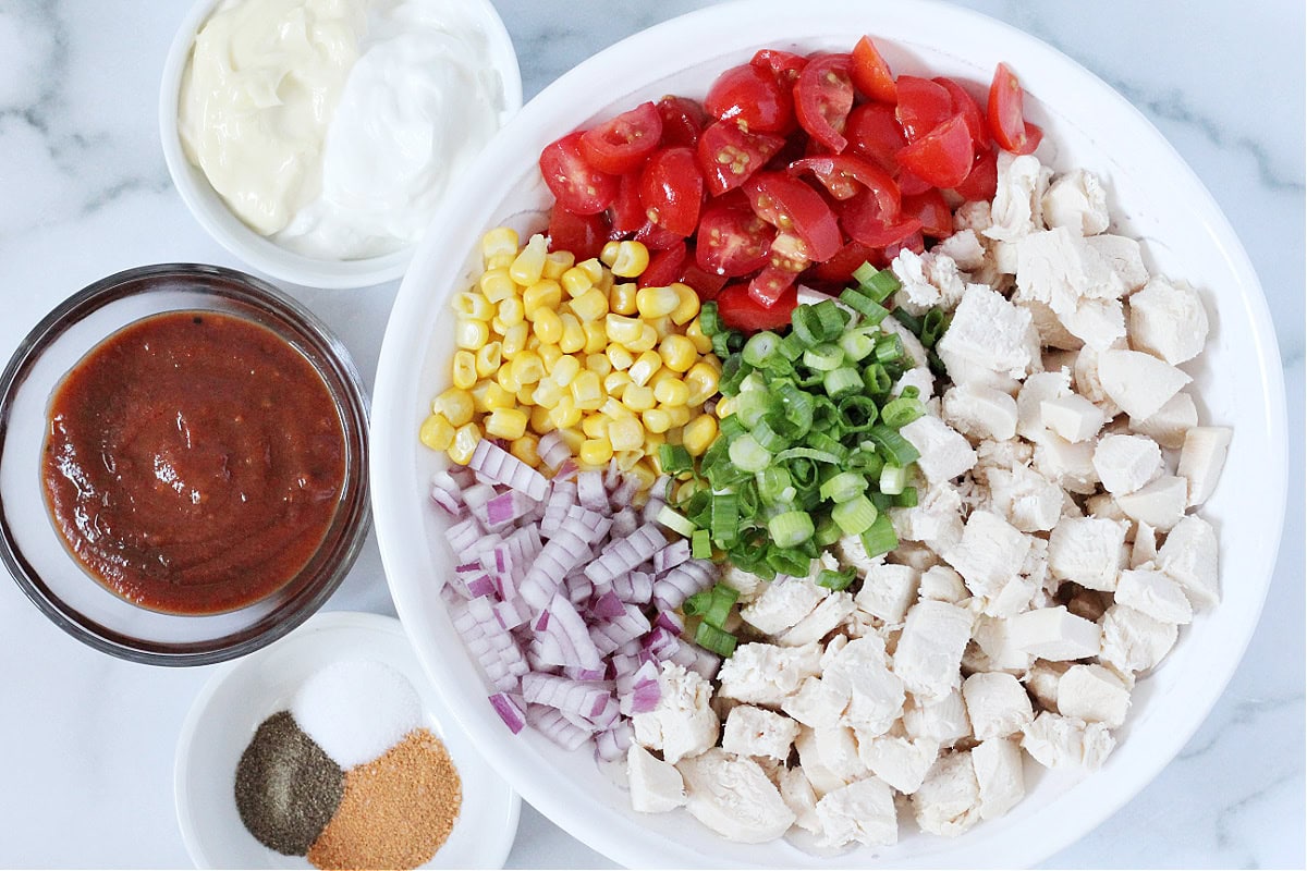 Diced chicken and fresh veggies in a white bowl with bowls of sauce and seasoning.