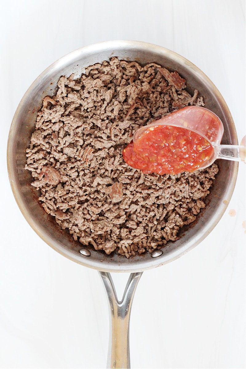 Salsa being poured into a pan of browned ground beef.