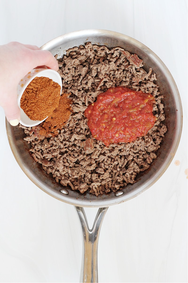 A woman's hand pouring taco seasoning into a metal pan with salsa and ground beef.