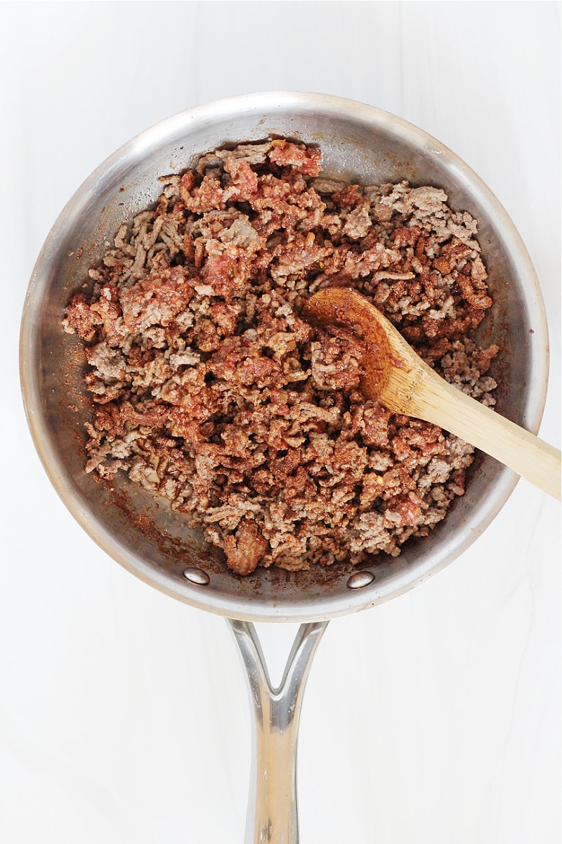 A wooden spoon stirring taco seasoning and salsa into browned ground beef.