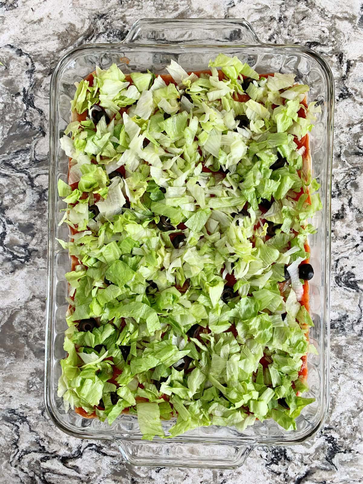 Overhead photo of chopped lettuce over black olives in a clear 9x13 pan.