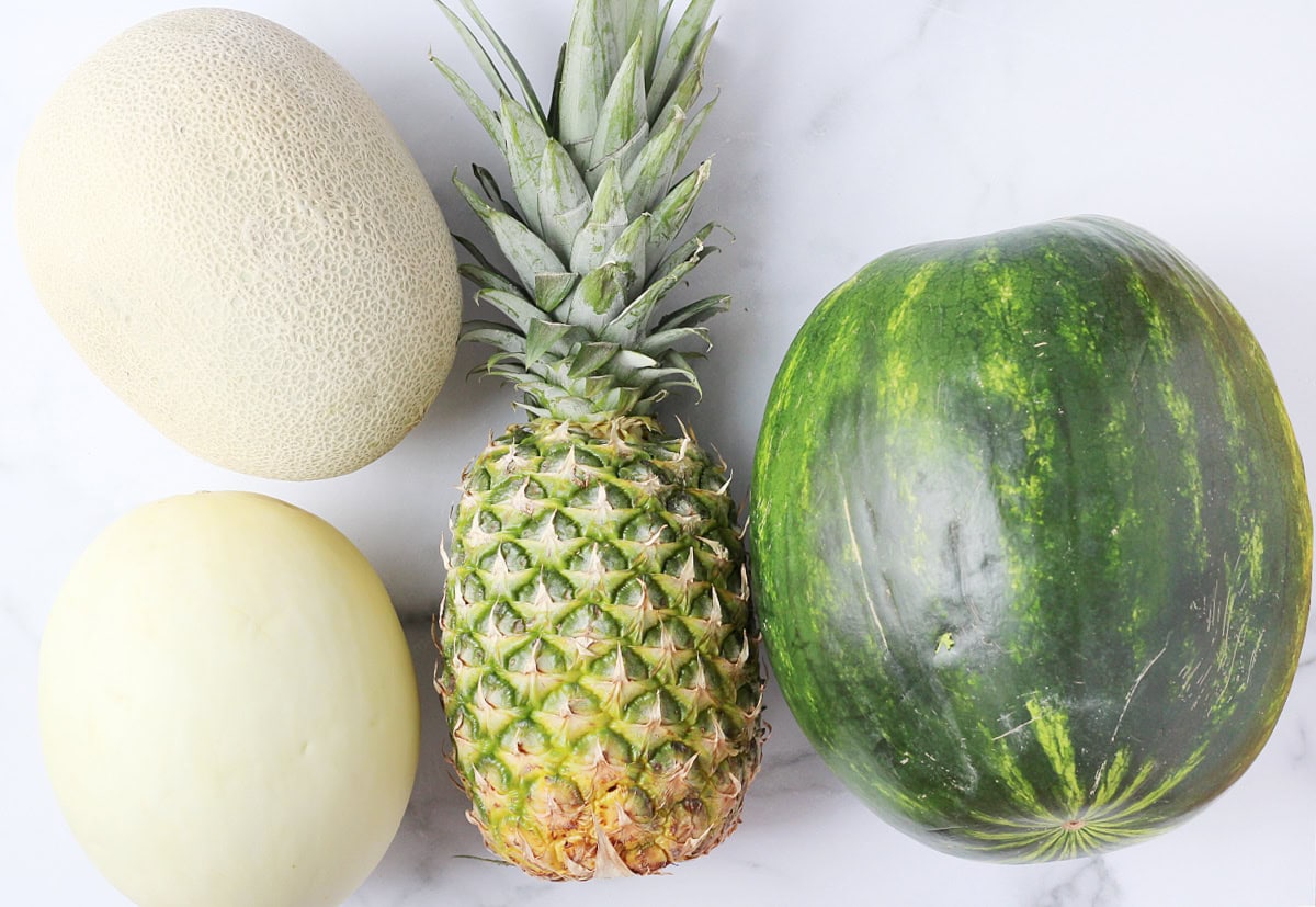 Overhead photo of a watermelon, pineapple, cantaloupe and honey dew melon on a white countertop.