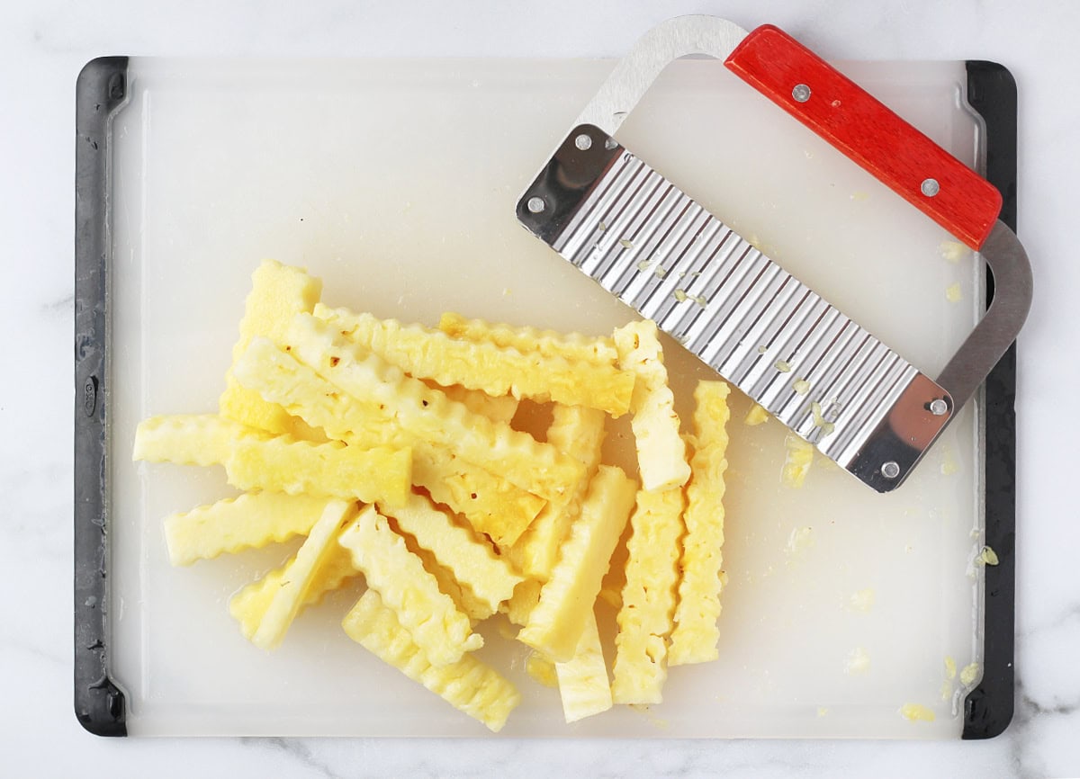 Overhead photo of pineapple fries and a metal crinkle cutter on a white cutting board.