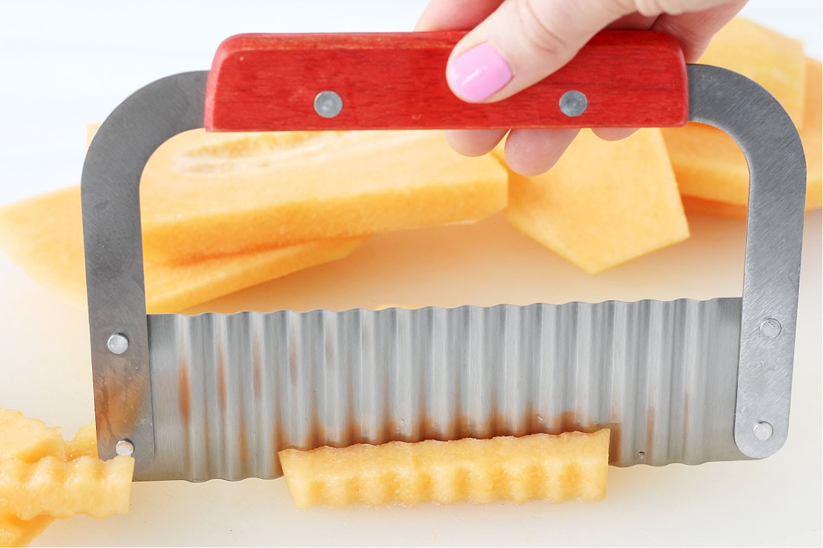 Photo of a metal crinkle cutter slicing cantaloupe fries on a white cutting board.