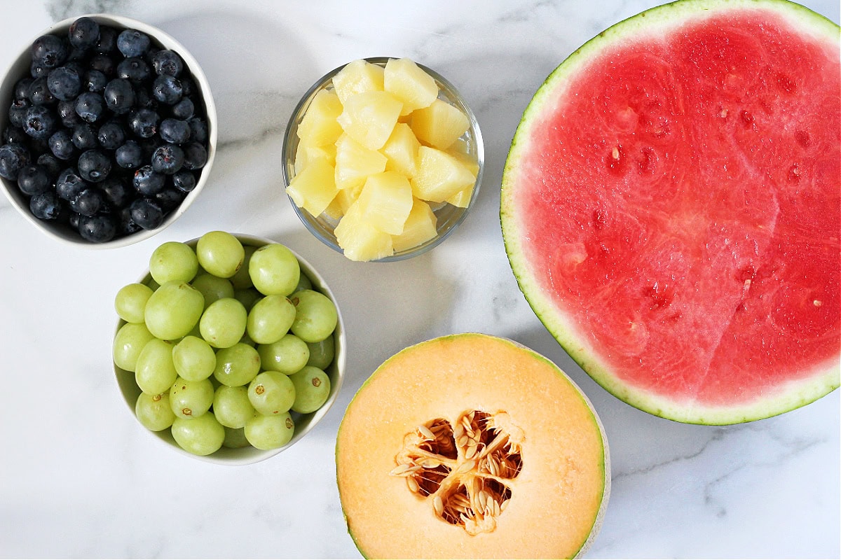 A halved watermelon and cantaloupe with bowls of diced pineapple, green grapes and blueberries.