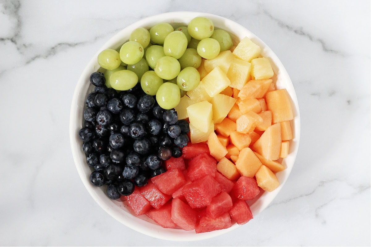 Fresh grapes, blueberries, pineapple, watermelon and cantaloupe separated in a white bowl.