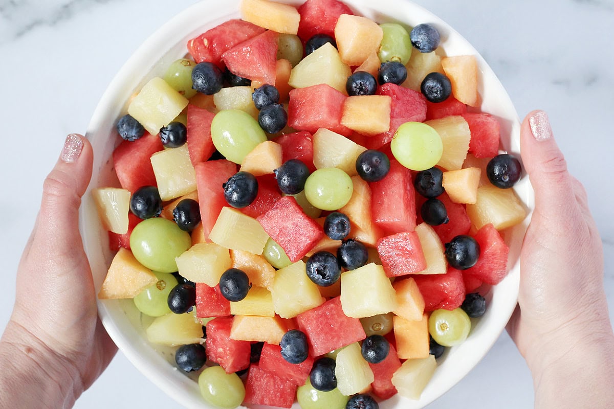 A woman's hands holding a white bowl of 5 ingredient fresh fruit salad.