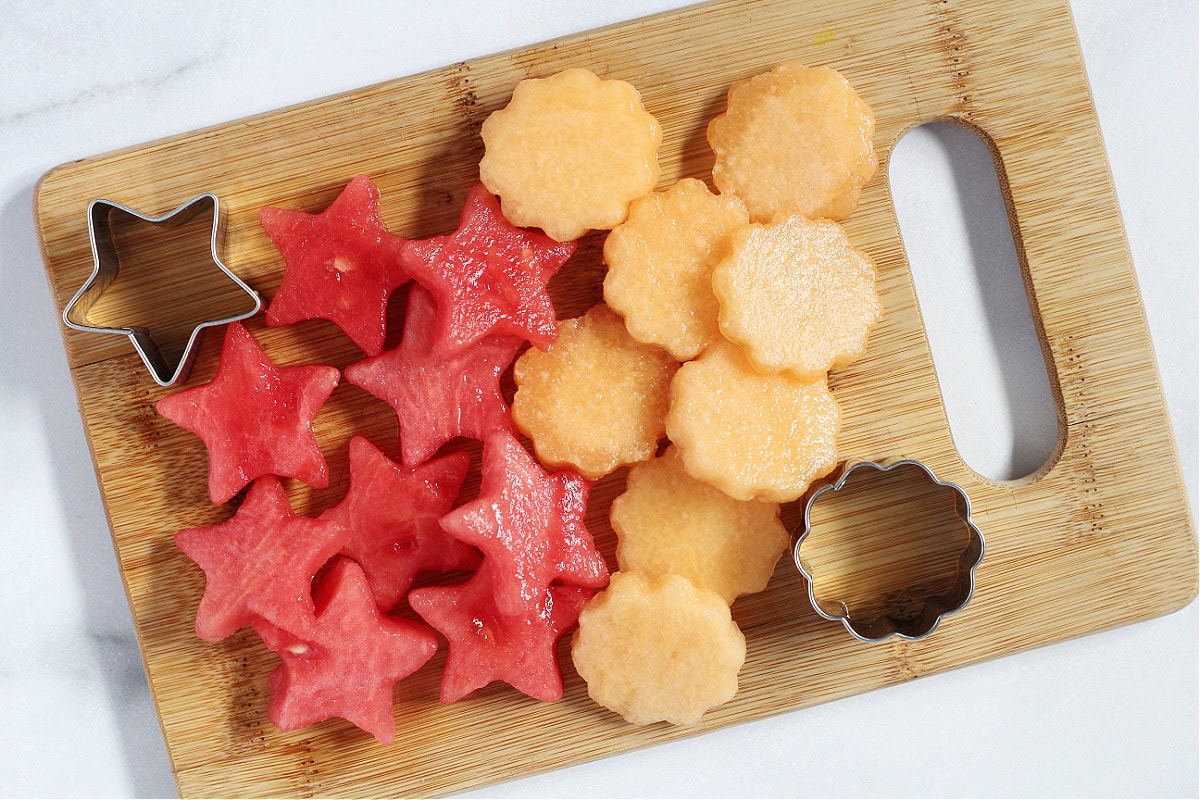 A wooden cutting board with star cutouts of watermelon and flower cutouts of cantaloupe.