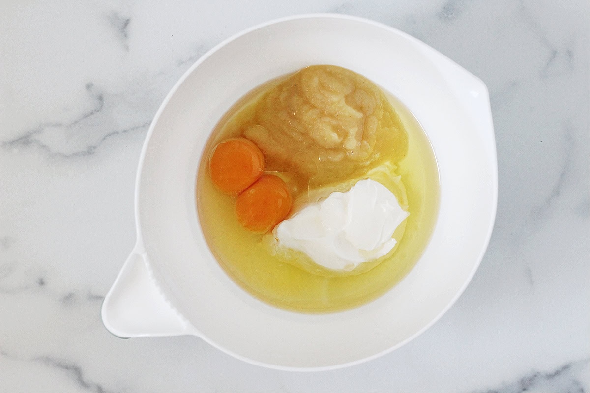 Overhead photo of eggs, sour cream, apple sauce and oil a white mixing bowl.