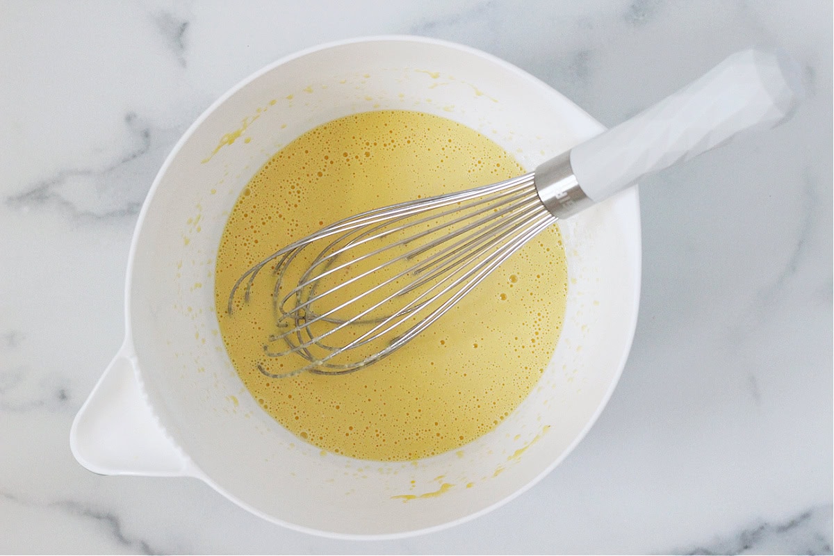 A white whisk in a bowl of eggs, oil, sour cream and mixed together.