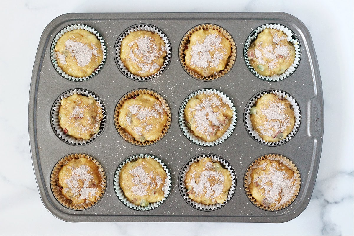 Overhead photo of twelve muffin cups with liners filled with rhubarb batter and cinnamon topping.