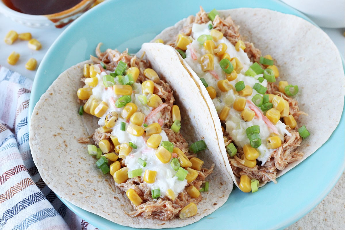 Two BBQ chicken tacos with coleslaw topped with corn on a blue plate.