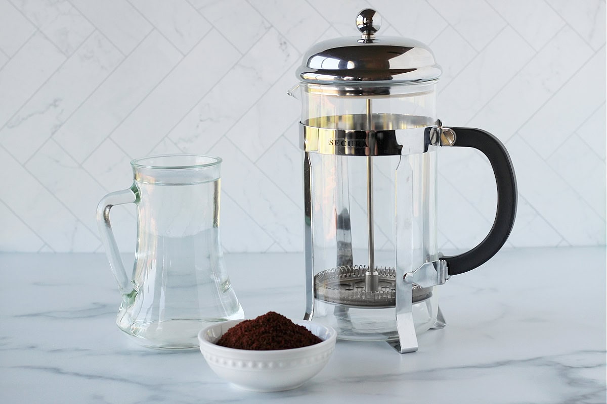 A water carafe, glass French press and a bowl of coffee grounds on a countertop.