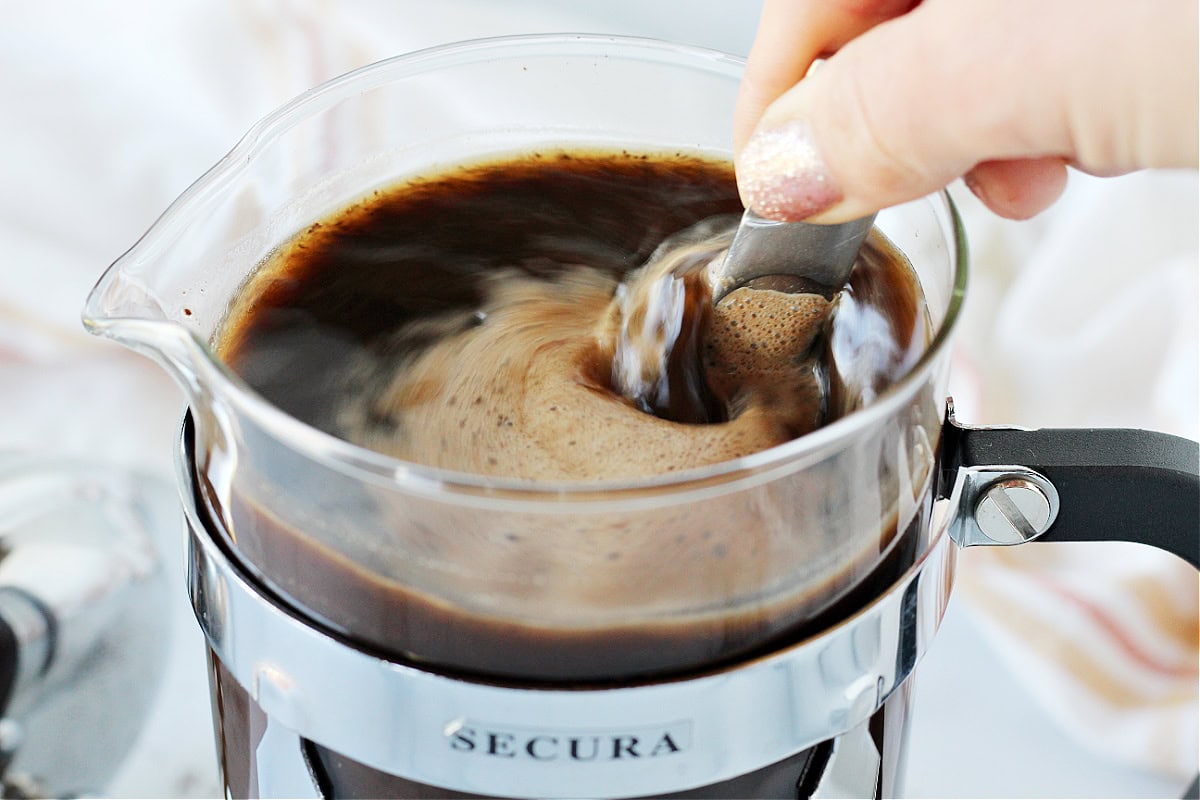 A spoon stirring coffee grounds and cold water in a French press.