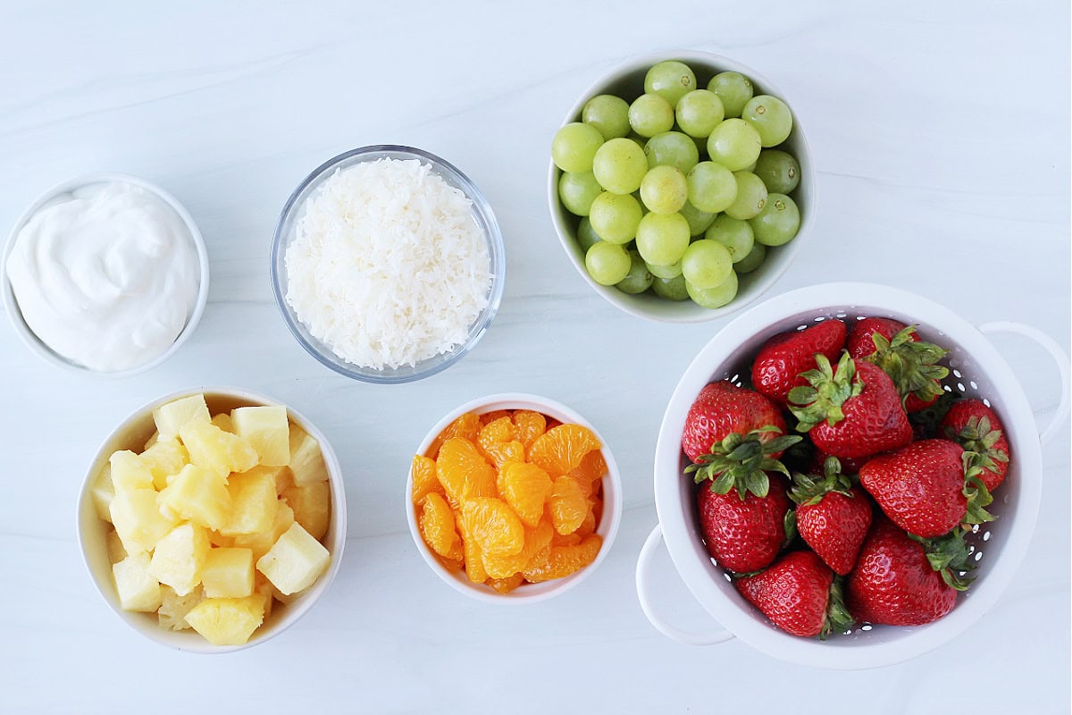 Bowls of ingredients for a coconut Hawaiian fruit salad on a white countertop.