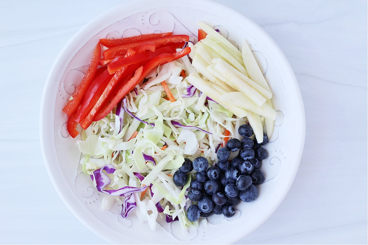 Julienned red bell peppers, blueberries, apple sticks, and coleslaw mix in a white bowl.