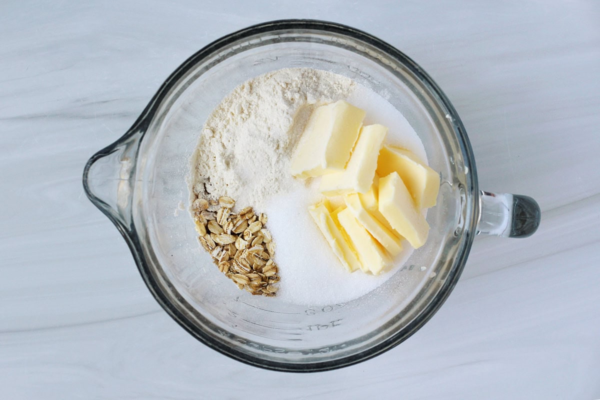 Rolled oats, flour, sugar and cubed butter ready to be combined in a glass bowl.