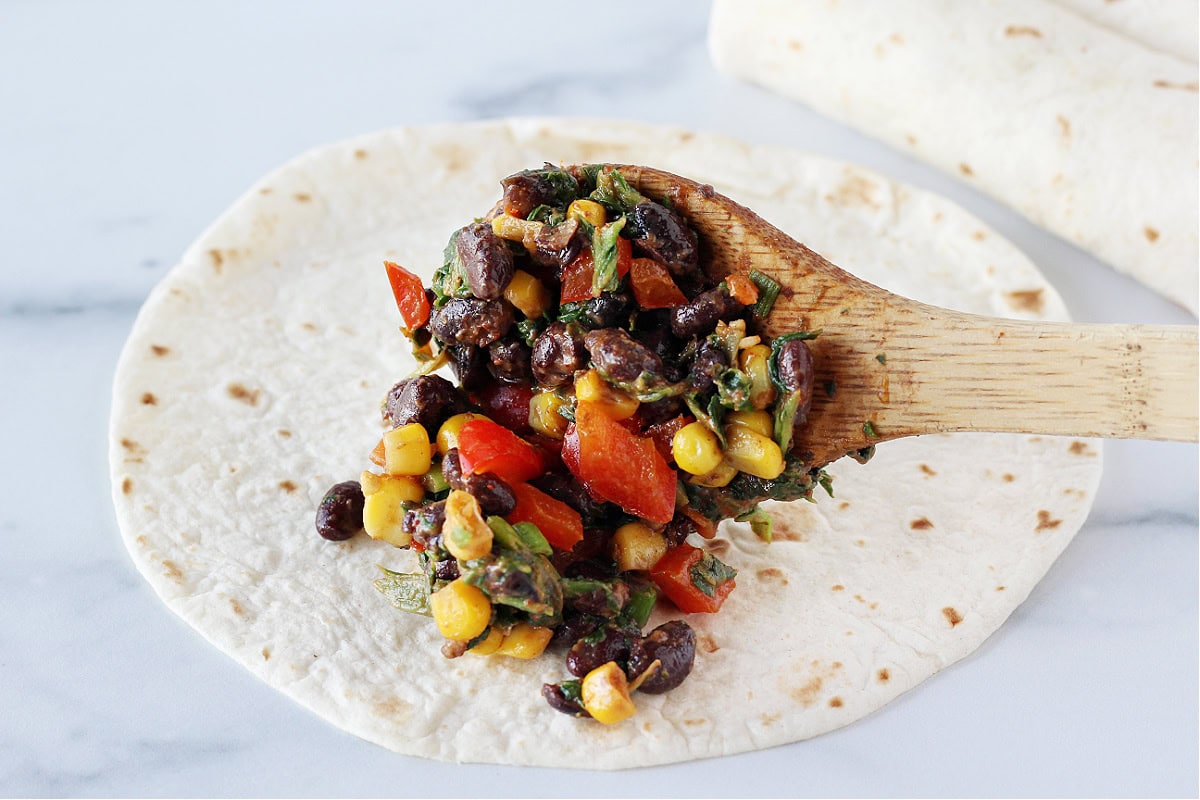 A wooden spoon putting a black bean filling on top of a flour tortilla.