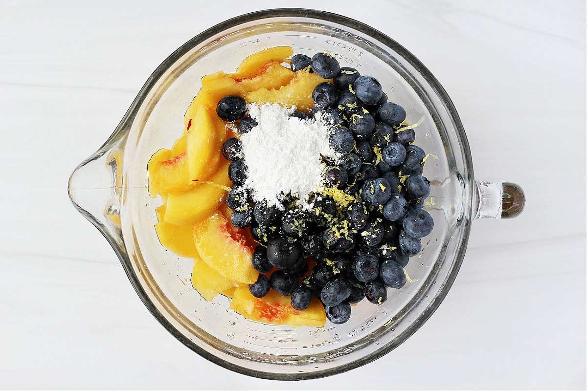 Overhead photo of sliced peaches, fresh blueberries, cornstarch, and lemon zest in a glass bowl.