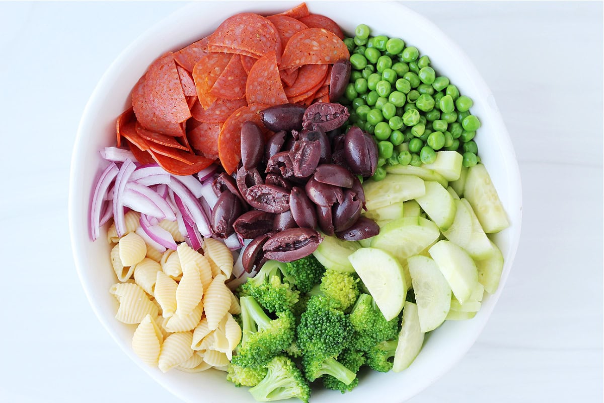 Overhead photo of sliced pepperoni, sliced vegetables, Kalamata olives and pasta shells in a bowl.