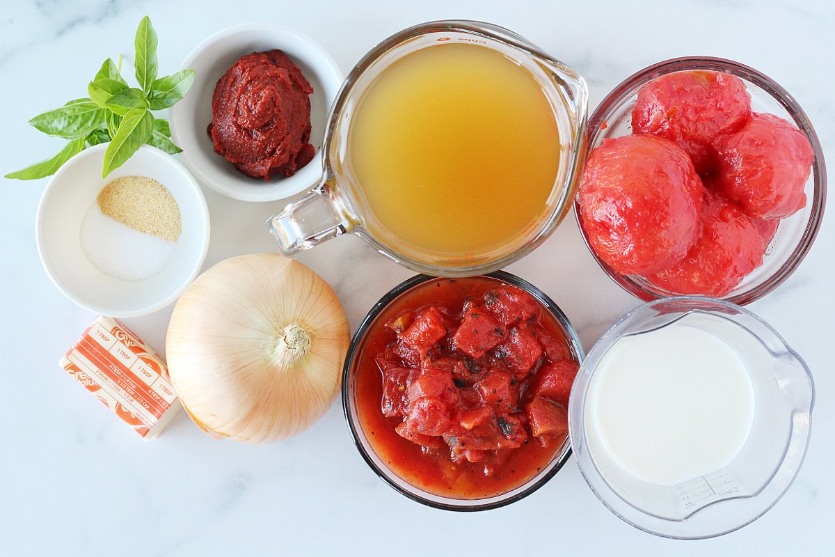 Ingredients for creamy homemade tomato soup with basil on a white marble countertop.