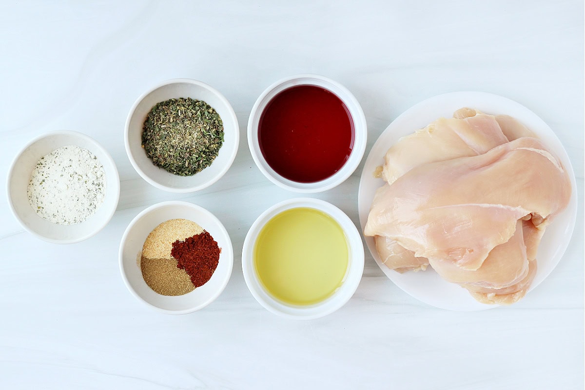 Raw chicken breasts on a plate with bowls of seasonings and sauces next to it.