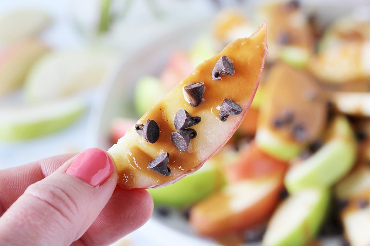 A woman's fingers holding up an apple slice dripping with peanut butter caramel sauce.