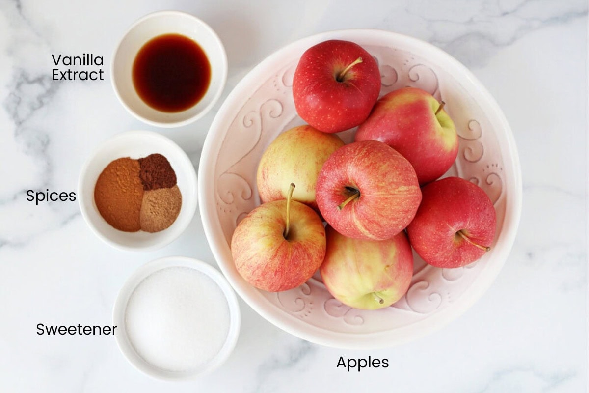 Red apples, vanilla extract, fall spices and sugar in white bowls on a marble countertop.