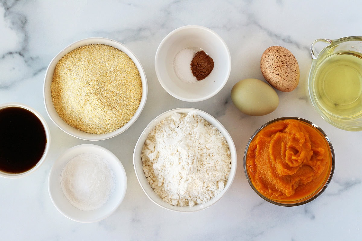 Ingredients for pumpkin cornbread in bowls on a white marble countertop.