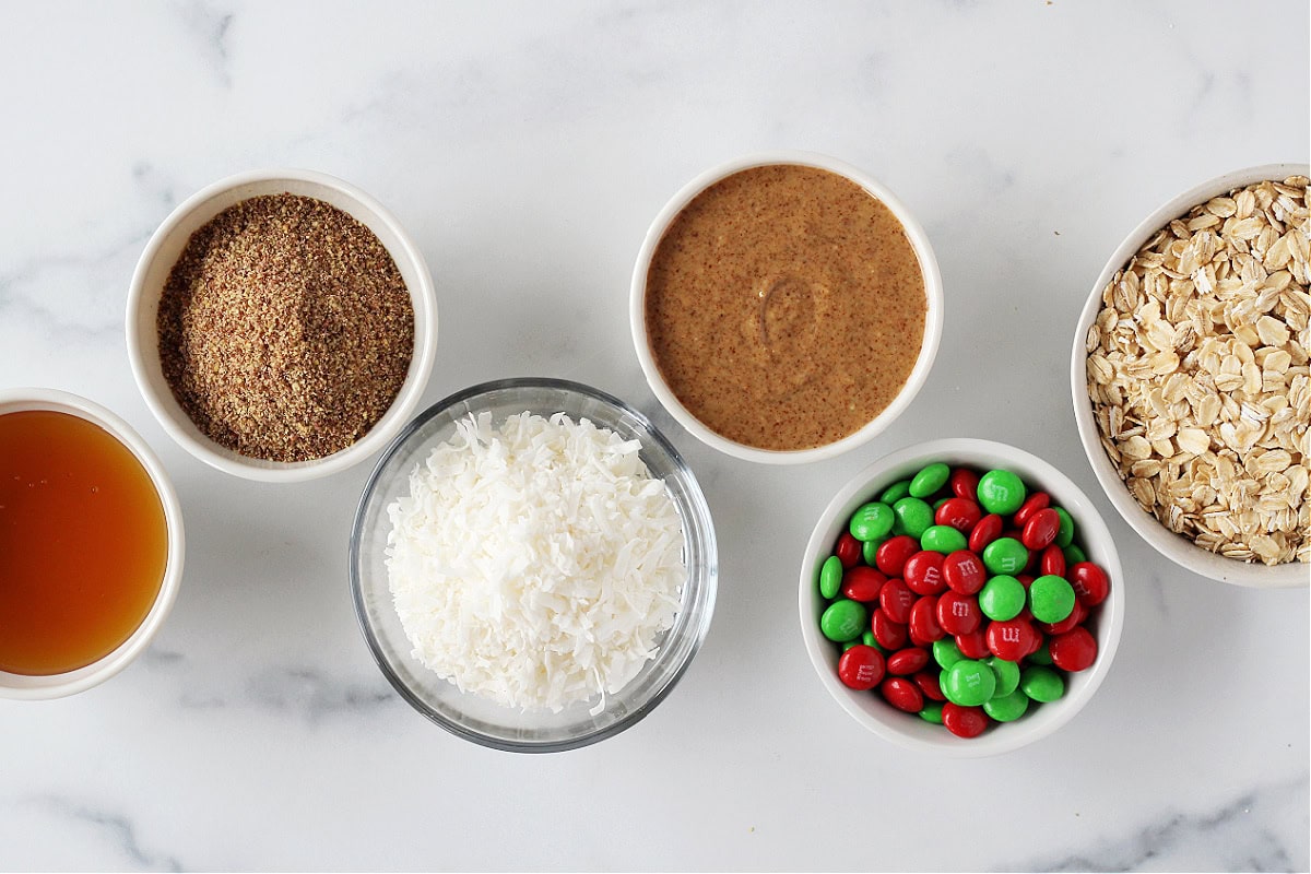 Bowls of ingredients for Christmas energy bites on a white marble countertop.