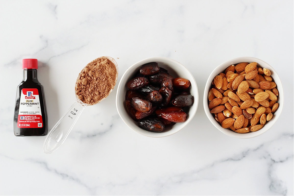 Peppermint extract, cocoa powder, dried dates and whole almonds on a white countertop.