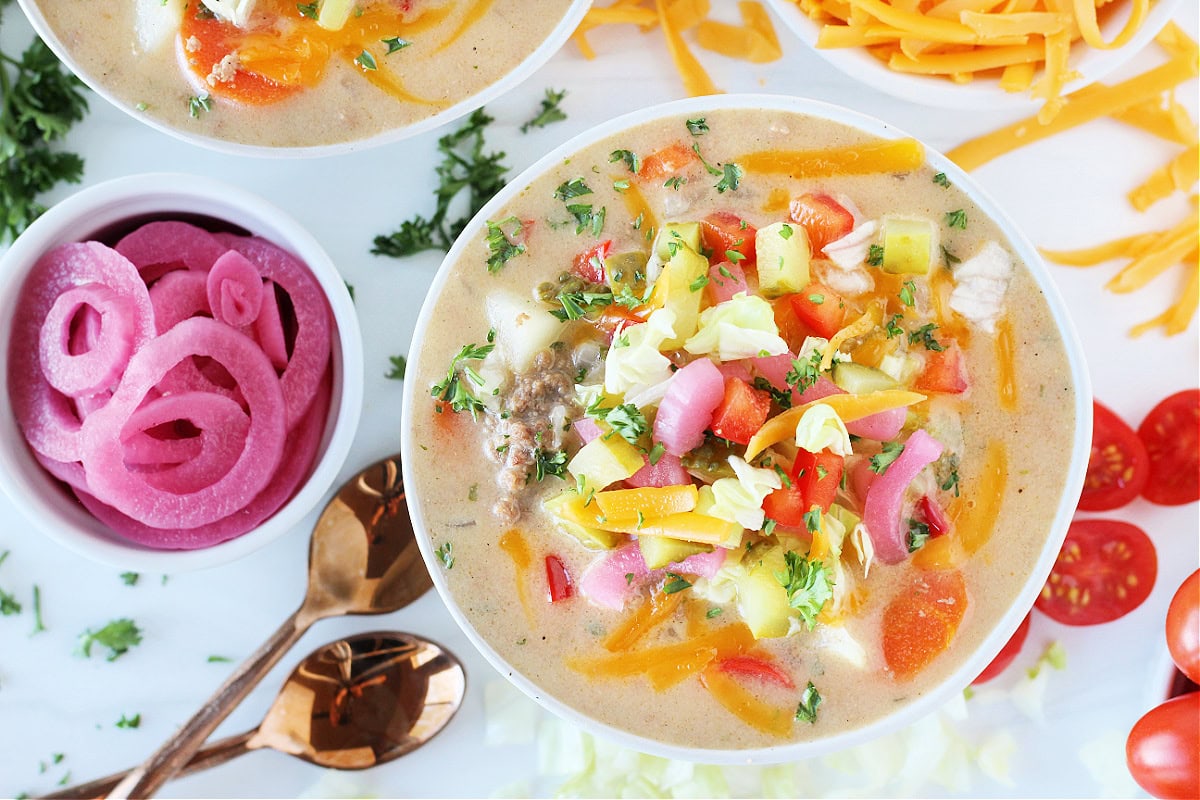 Overhead photo of a bowl of cheesy soup topped with pickled onions, pickles and cheese.