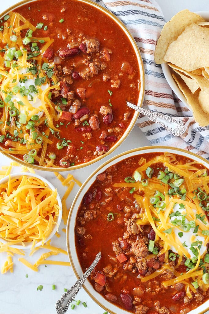 Overhead photo of two bowls of sweet and spicy chili with toppings and spoons.