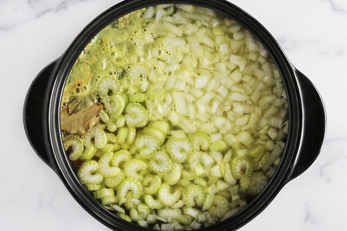 Diced onion, sliced celery, and bay leaves in vegetable broth in a black pot.