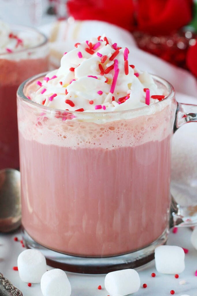 Red velvet hot cocoa in a glass mug topped with whipped cream and sprinkles.