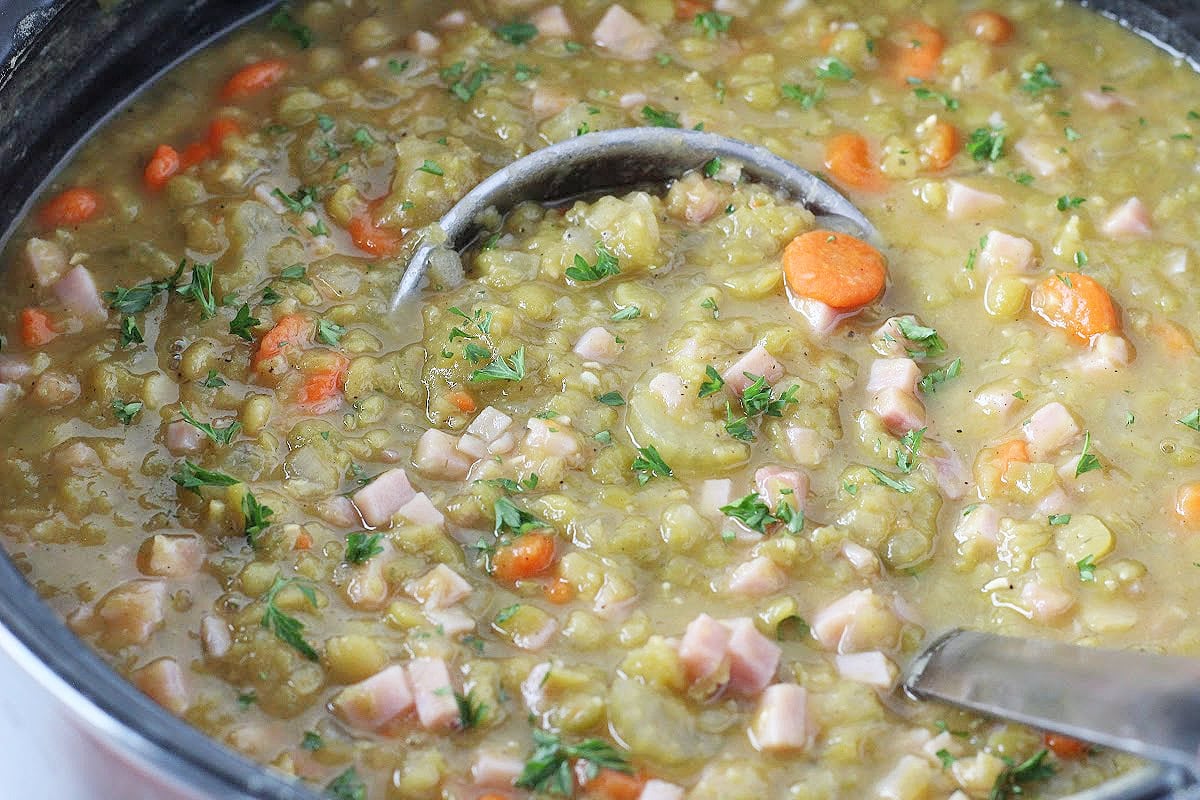 A metal ladle in a pot of split pea soup with ham and carrots.