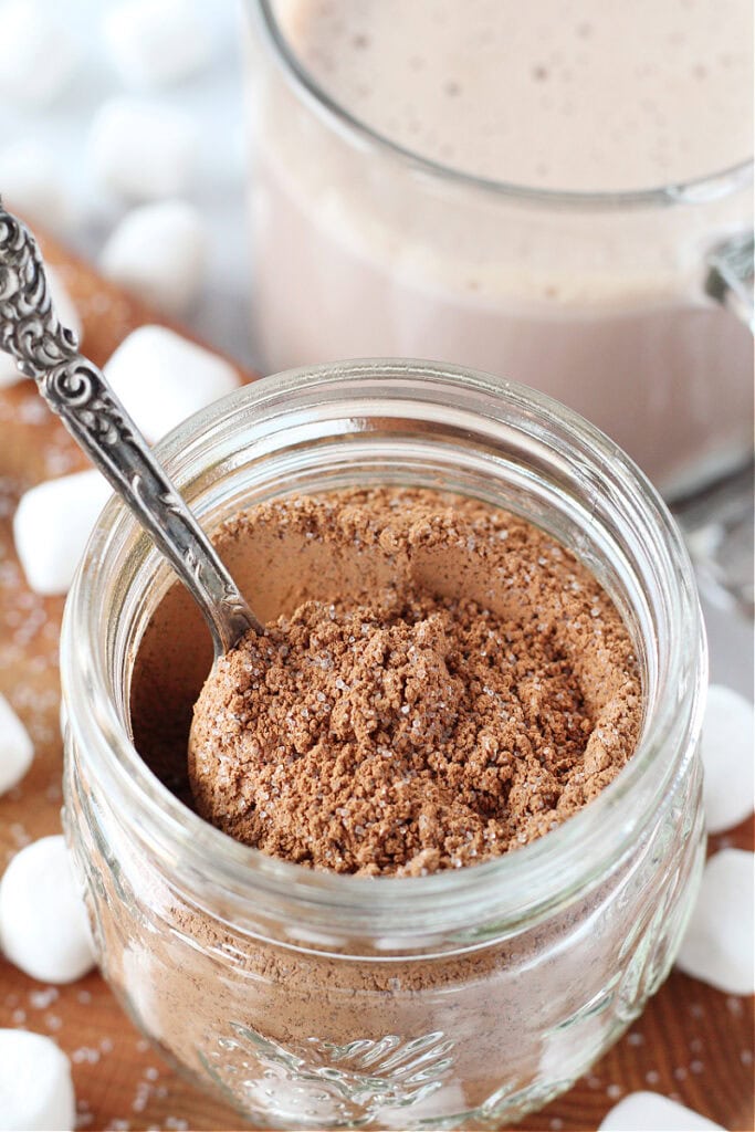 Clear jar filled with hot cocoa mix and a spoon with mug of hot chocolate.