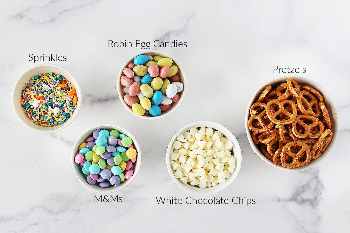 Labeled ingredients for white chocolate pretzel bark with Easter candies on a white marble countertop.