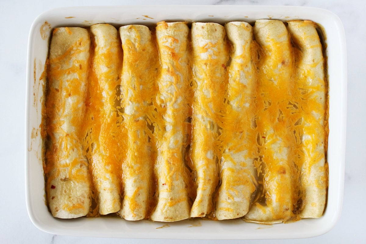 Eight baked enchiladas topped with melted cheese in a white baking dish.