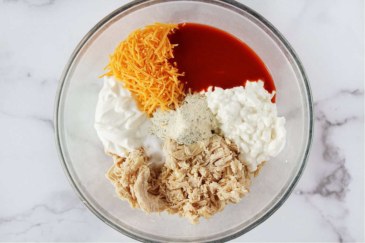 Buffalo sauce, cottage cheese, chicken and shredded cheese in a glass bowl.