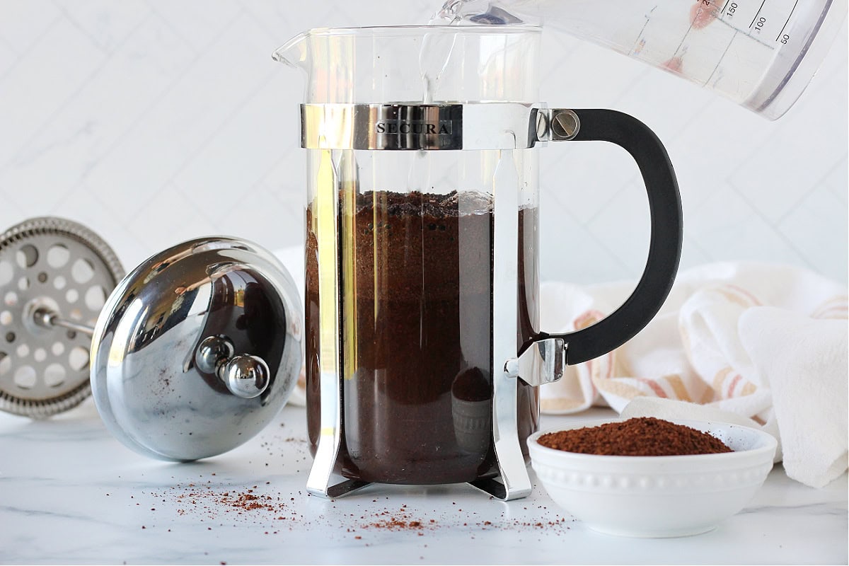Water pouring into a glass French press with coffee grounds.