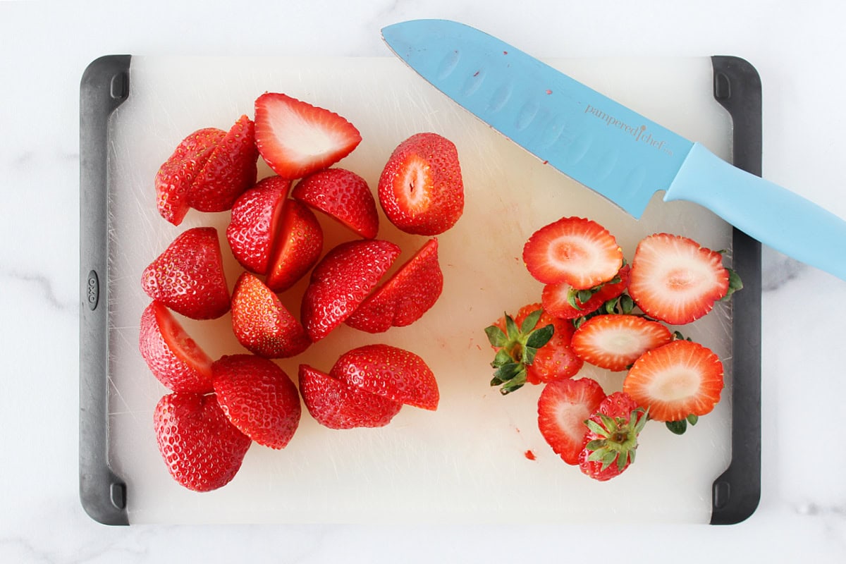 Fresh strawberries with the tops removed and sliced in half on a cutting board.