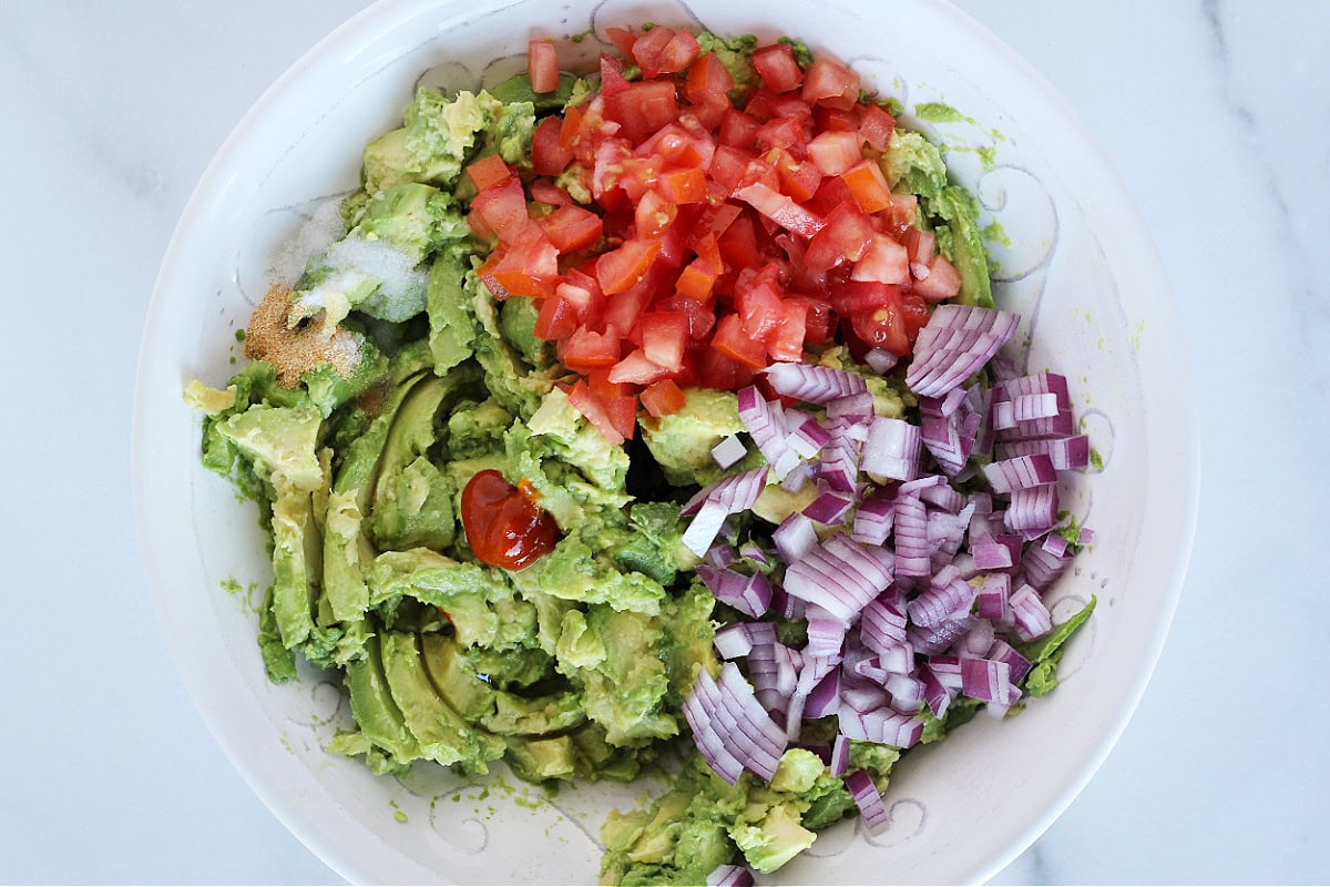 Chopped veggies, seasonings, sriracha sauce and mashed avocados in a bowl.
