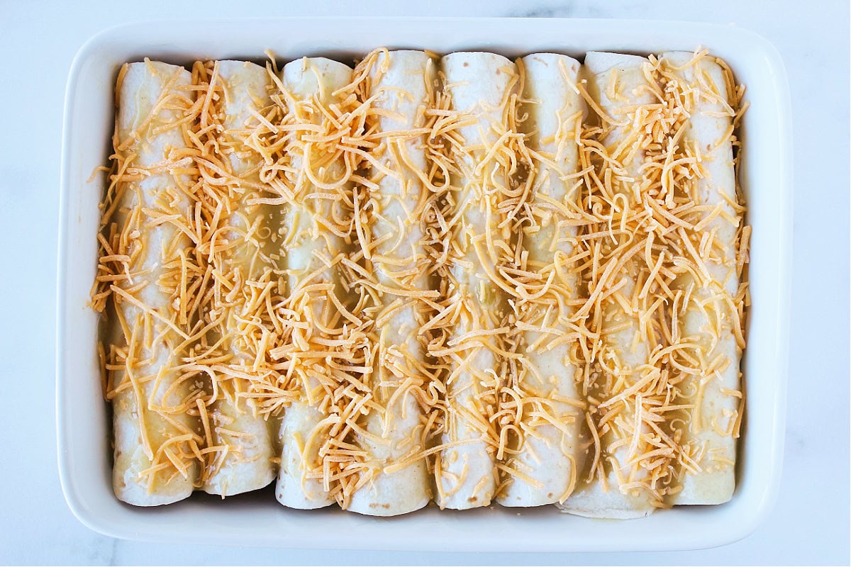 Unbaked enchiladas topped with green enchilada sauce and shredded cheese.