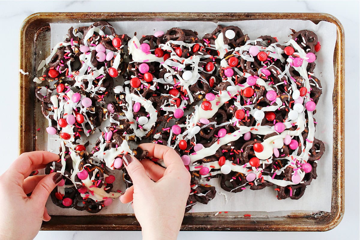 A woman's hands breaking apart Valentine's Day chocolate pretzel bark on a sheet pan.