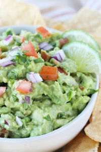 Guacamole in a white bowl topped with chopped red onion, tomato, and cilantro.